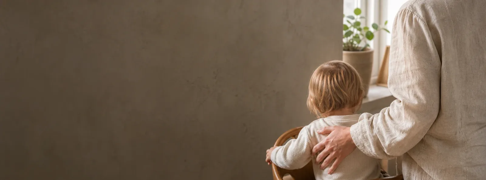 A parent settles a young child at the family table in soft morning light.